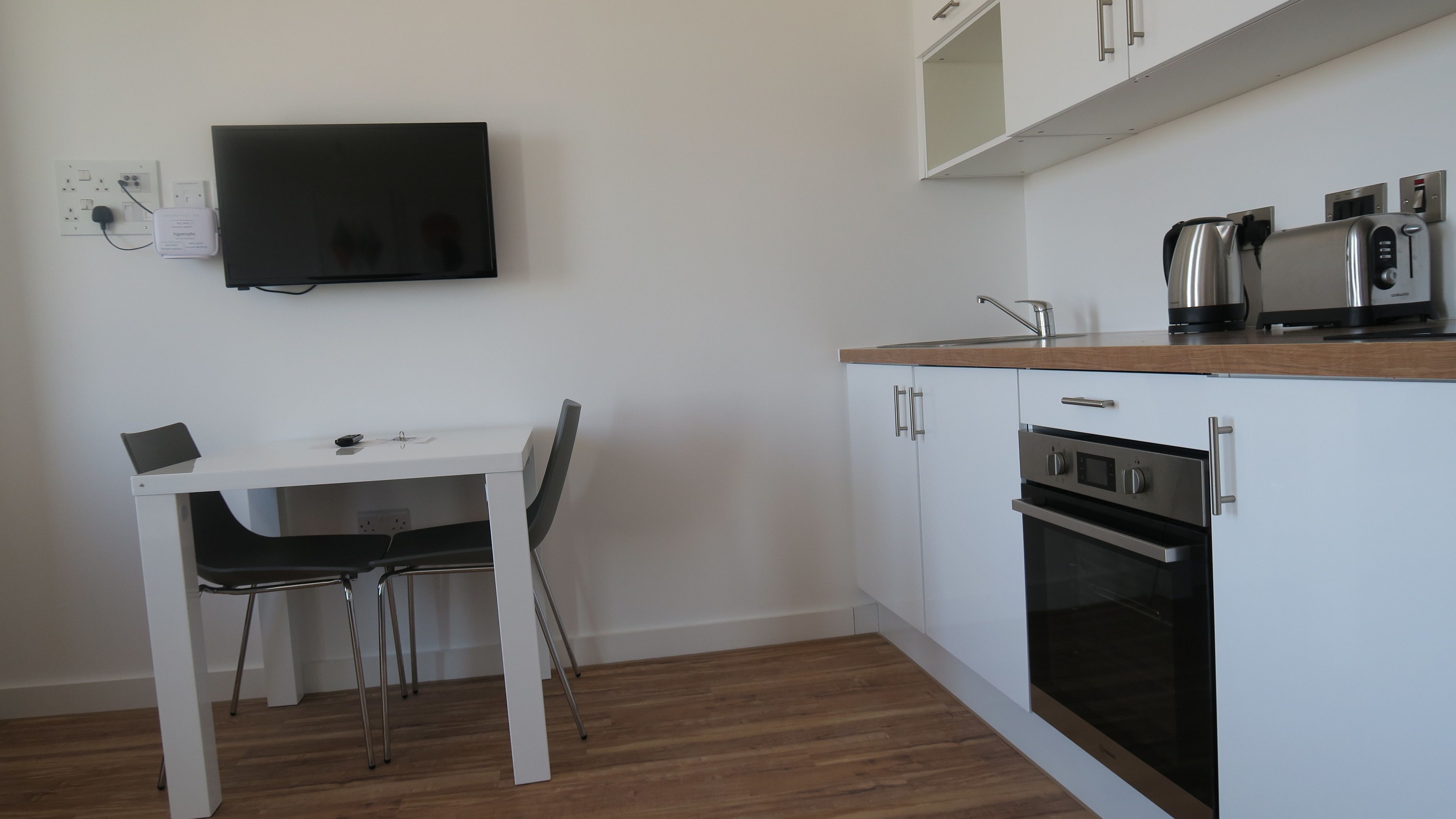 Una cocina compacta con armarios blancos, horno empotrado, cafetera y televisor de pantalla plana montado en la pared.