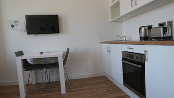 Una cocina compacta con armarios blancos, horno empotrado, cafetera y televisor de pantalla plana montado en la pared.