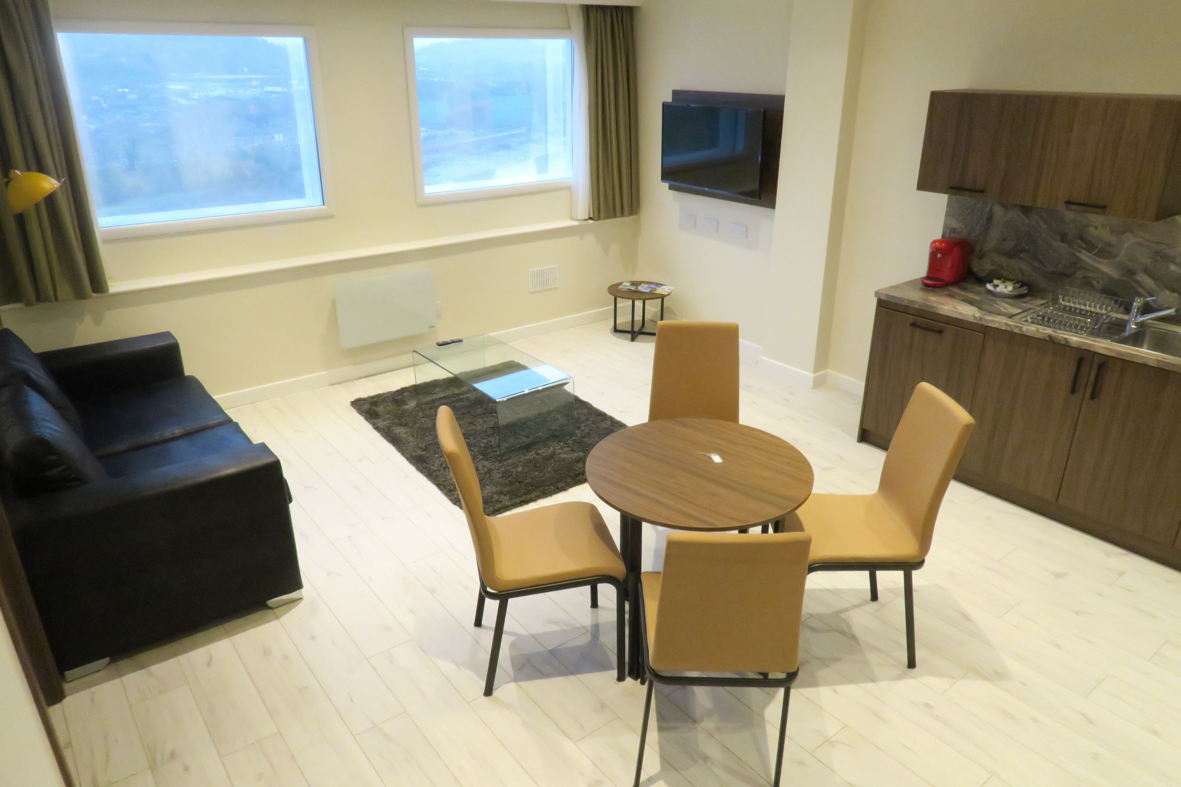 A modern living room with a black sofa, a glass coffee table, and a round wooden table with four chairs. There are two windows with curtains, a flat-screen TV mounted on the wall, and a small side table with a red object.