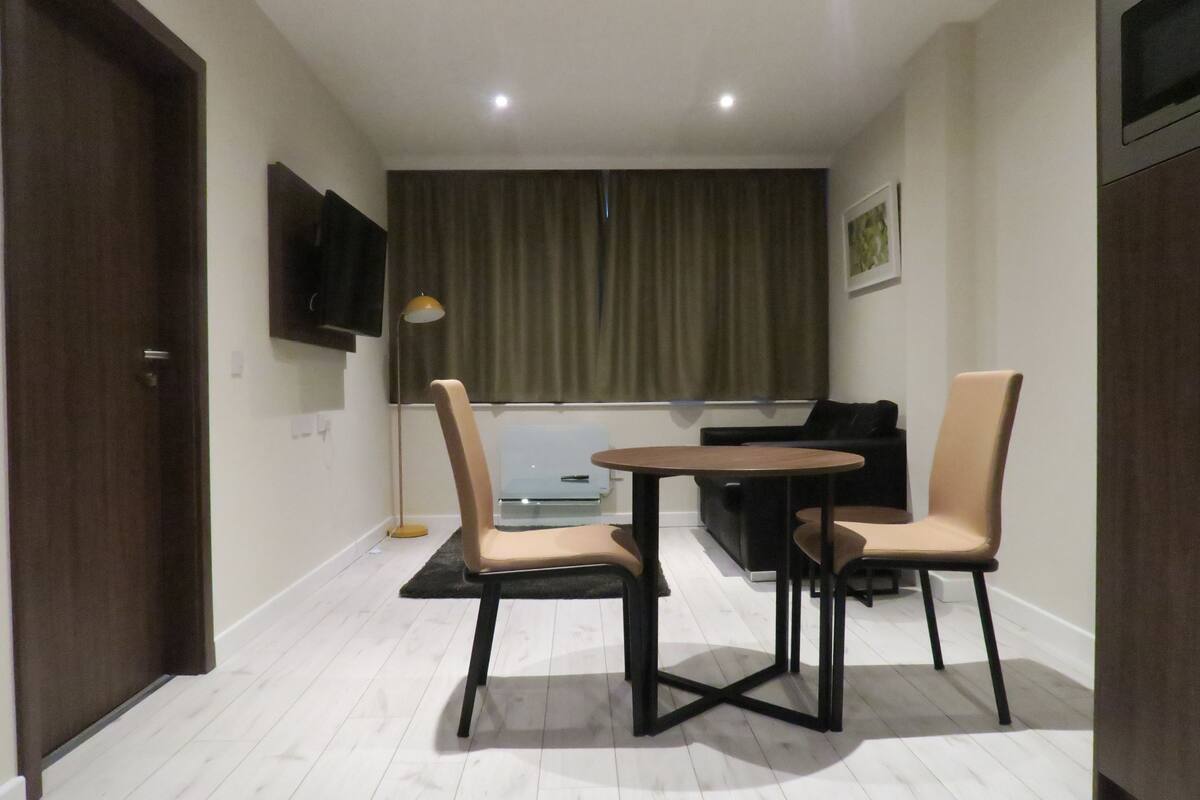 A small room with a dining table and two chairs, a TV mounted on the wall, and a window with curtains.