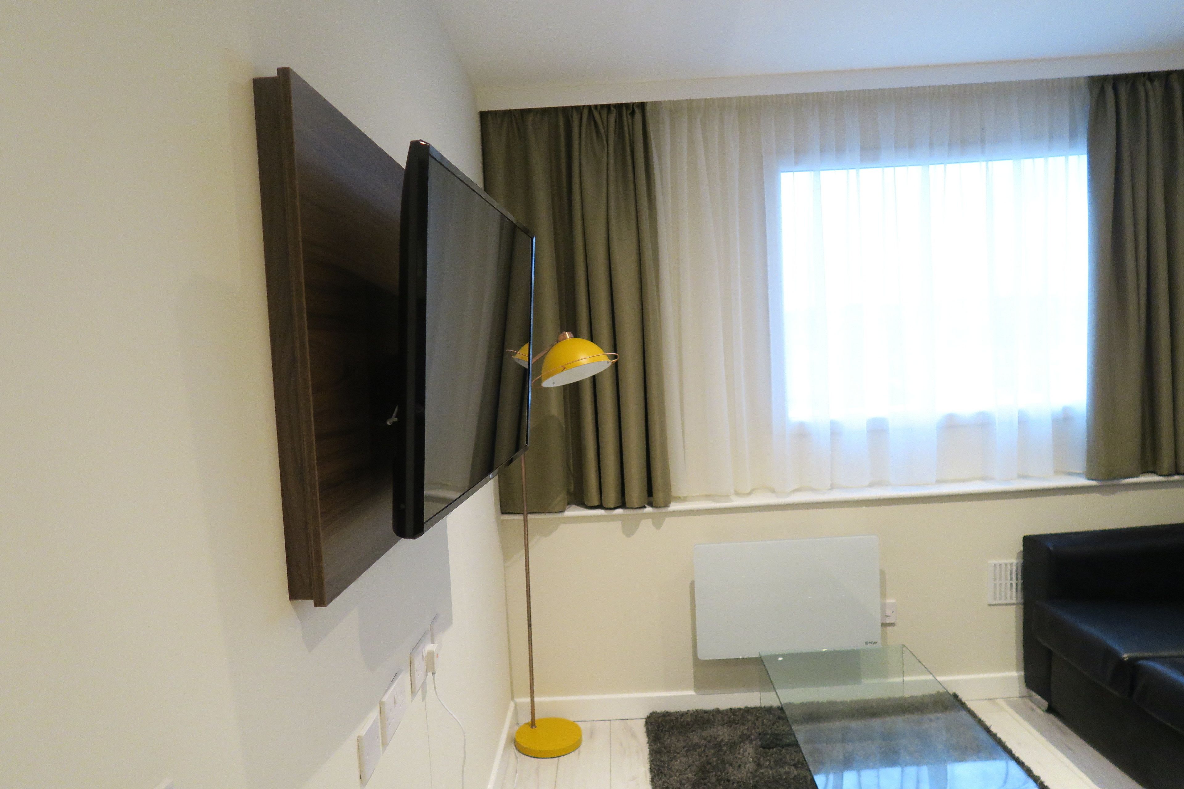 Un salon moderne avec un téléviseur à écran plat fixé au mur, une table basse en verre, un canapé noir et une lampe de sol jaune.