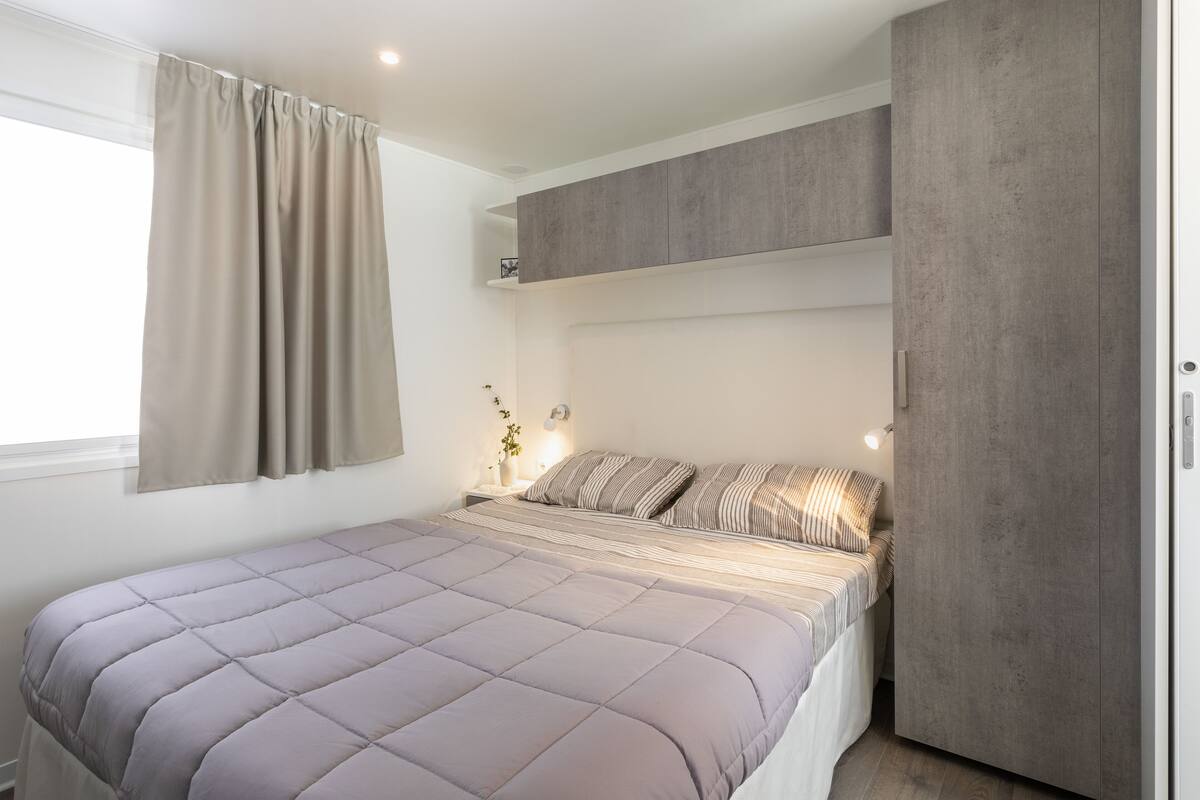 A bedroom with a large bed, a wardrobe, a bedside table with a lamp, and a window with curtains.