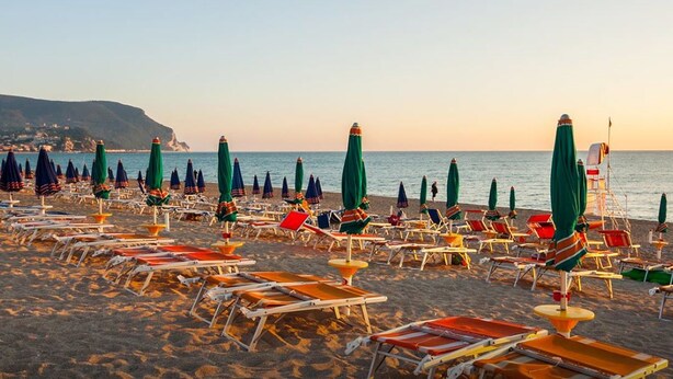 A beach with rows of sun loungers and umbrellas, a boat in the water, and a coastline in the background. - Camping Village Riviera (Numana)