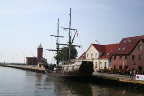 A historic sailing ship docked at a waterfront, with a red-roofed building and a lighthouse in the background.