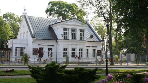 A white building with a steeply pitched roof, large windows, and a symmetrical facade. - Villa Karmelia (Darlowo)