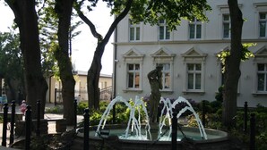 A fountain with a statue in the center, surrounded by trees and a building with multiple windows in the background. - Villa Karmelia (Darlowo)