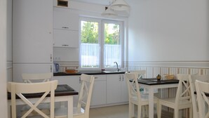 A bright kitchen with white cabinets, a black countertop, and white chairs. There are two windows with white curtains, and a white pendant light hangs above the counter. - Villa Karmelia (Darlowo)