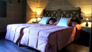 A bedroom with a large bed, two bedside lamps, and a stone wall. - Casa Rural Estrela (San Martin de Trevejo)
