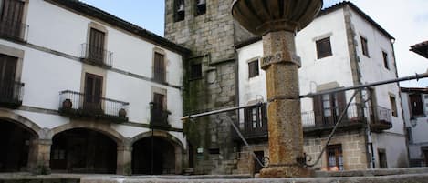 Una plaza histórica con una fuente central, edificios de piedra con balcones y una torre del reloj.