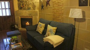A cozy living room with a stone fireplace, a dark blue sofa with floral cushions, a glass coffee table with books, and a star decoration on the wall. - Casa Rural Estrela (San Martin de Trevejo)