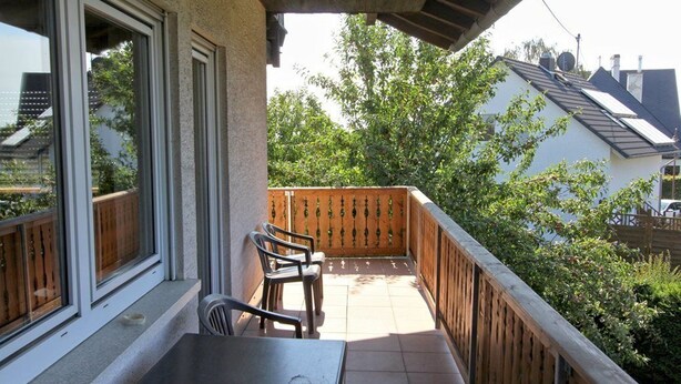 Een balkon met een tafel en stoelen, houten reling en uitzicht op een huis en bomen.