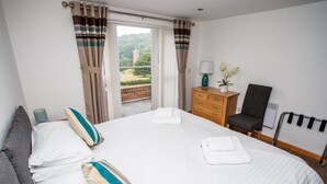 Habitación de hotel con una cama grande, un aparador de madera, una silla y vistas a un edificio y vegetación por la ventana.