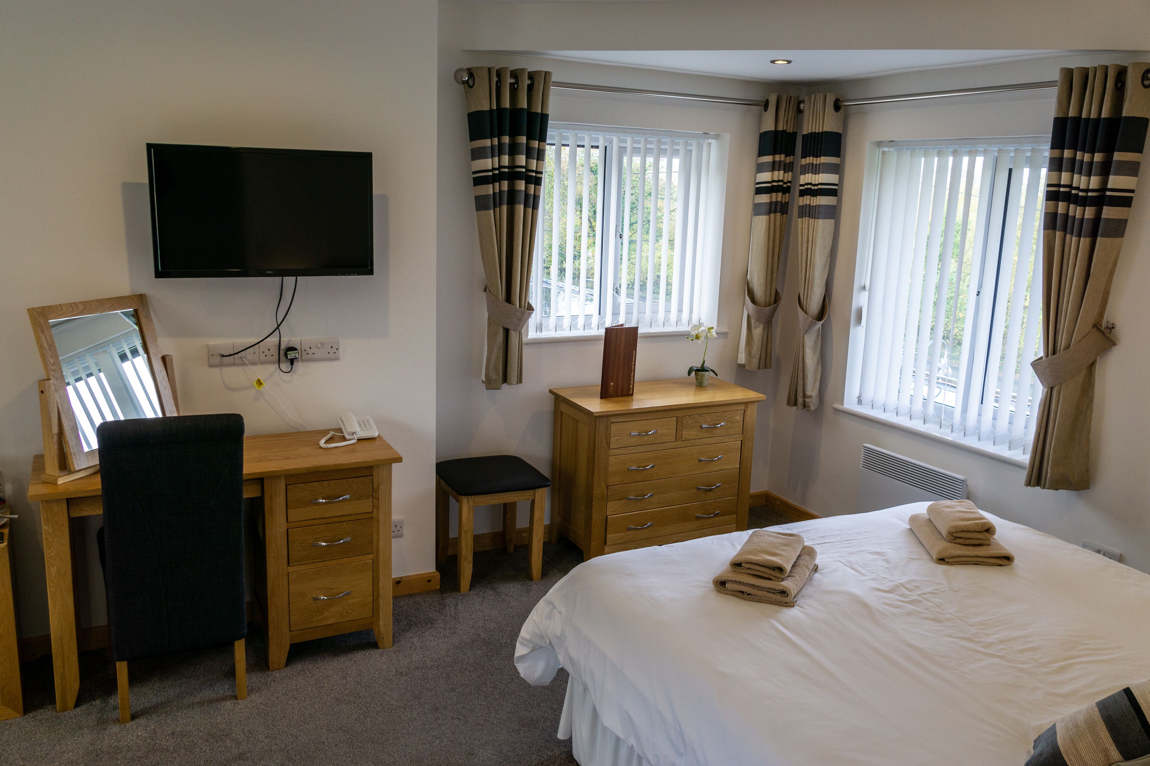 A hotel room with a bed, a desk with a chair, a dresser, a TV mounted on the wall, and a window with curtains.