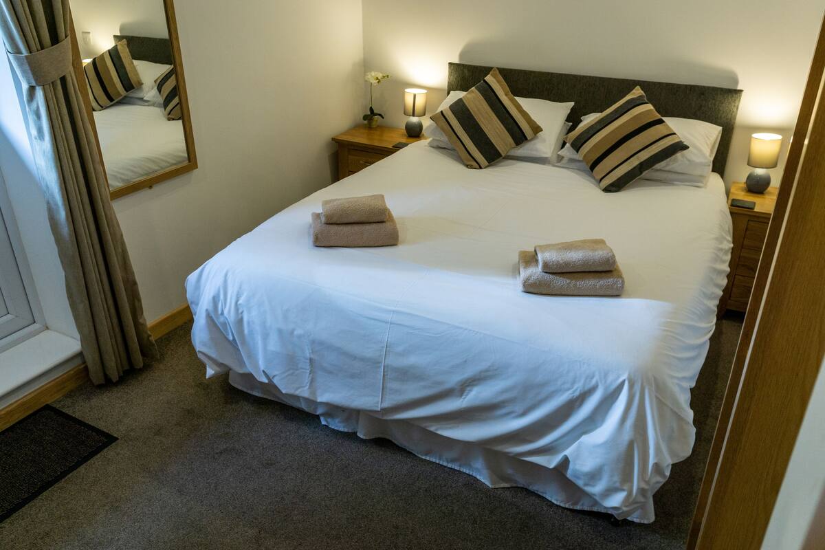 A neatly made bed with white linens and striped pillows, a bedside table with a lamp, and a mirror on the wall.