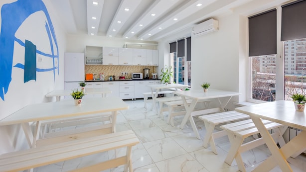 Una cocina moderna con armarios blancos, un microondas y un lavabo. Hay mesas y bancos blancos, y un ventanal con vistas a edificios.
