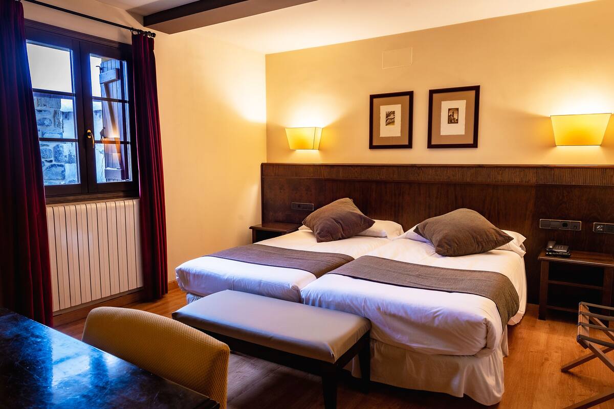 A hotel room with two beds, a bench, a table, and a window with curtains.