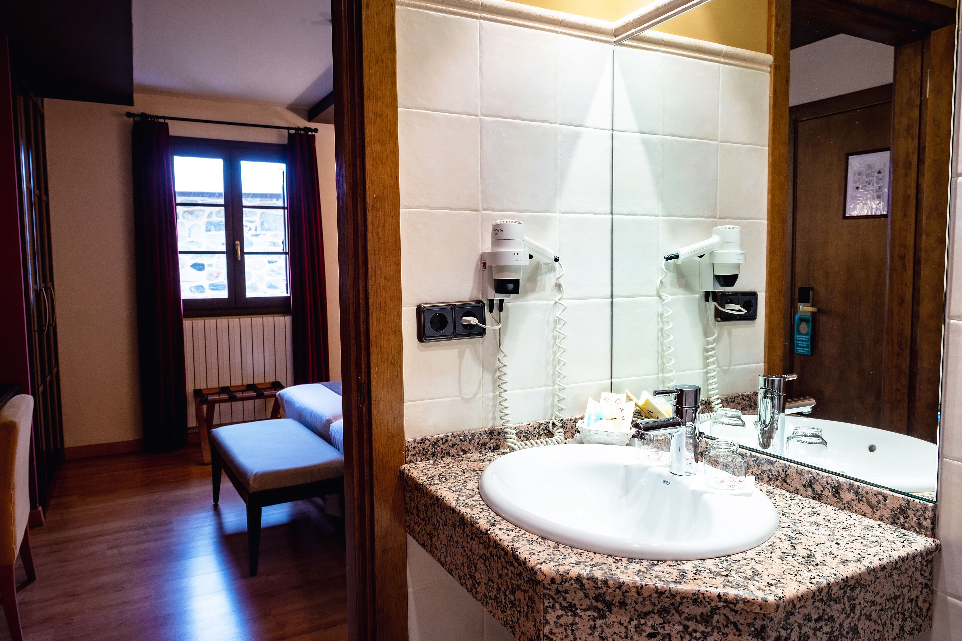 Ein Badezimmer mit einer Granit-Arbeitsplatte, einem weißen Waschbecken und einem großen Spiegel. Auf der Platte liegen ein Fön und eine Shampoo-Flasche.