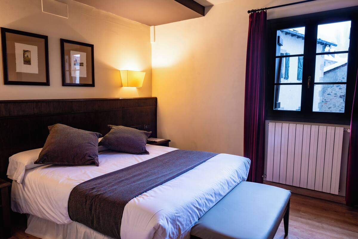 A hotel room with a large bed, a bedside table, a lamp, a window with curtains, and framed pictures on the wall.