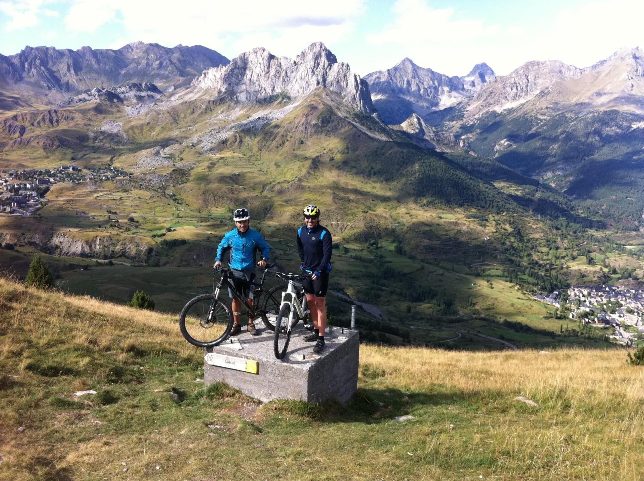 Due ciclisti in piedi su un monumento in pietra con un paesaggio montuoso sullo sfondo.