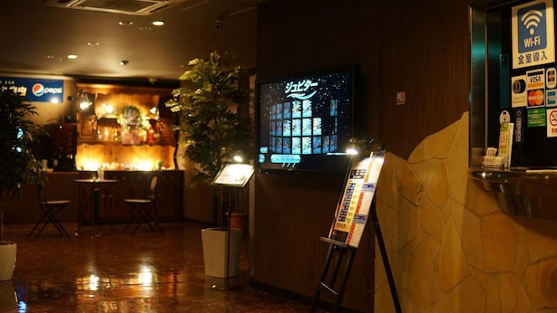 Un vestíbulo de hotel con una zona de bar, una pantalla digital que muestra un menú y un cartel informativo.