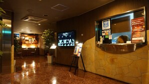 Un vestíbulo de hotel con una mesa de recepción, una pantalla digital que muestra un paisaje urbano, un atril con menús y una zona de estar con mesas y sillas.