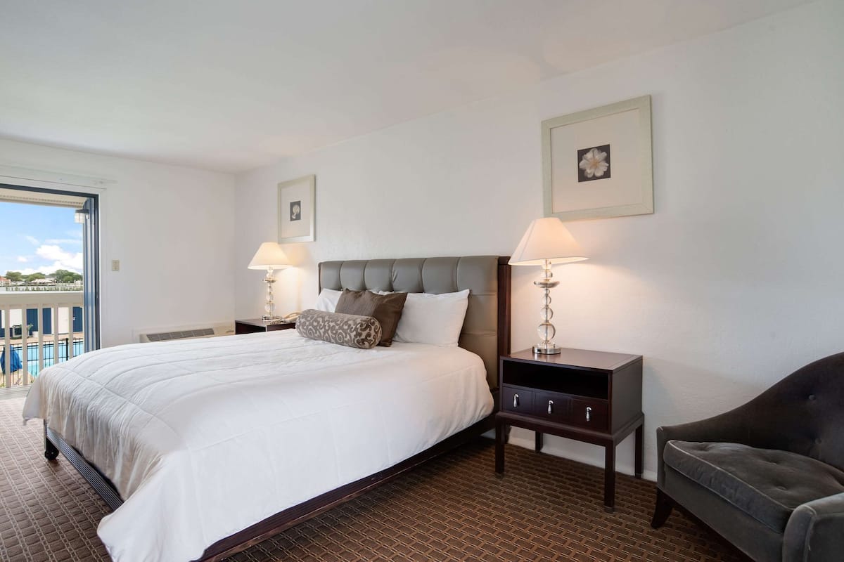 Habitación de hotel con una cama grande, dos mesitas de noche con lámparas, un sillón oscuro y vistas a una piscina a través de una puerta de cristal.
