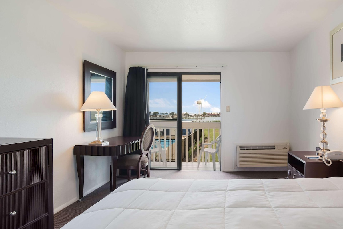 Habitación de hotel con cama, escritorio, silla, espejo, lámpara, teléfono y vista a una piscina y un edificio a través de una puerta de vidrio.