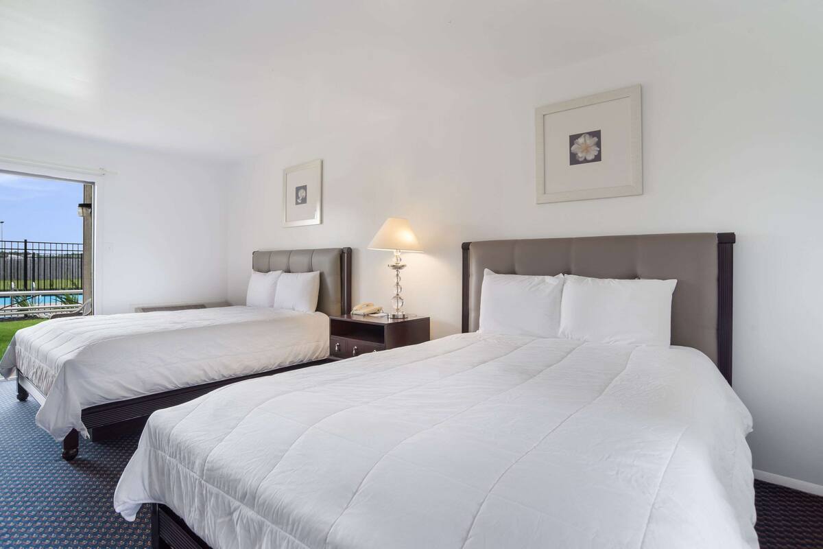 Habitación de hotel con dos camas, una mesita de noche con lámpara y una ventana con vistas al exterior.