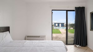 A hotel room with a bed, an air conditioning unit, and a sliding glass door leading to a balcony with a pool and greenery. - Rodeway Inn at Somers Cove Marina (Crisfield)