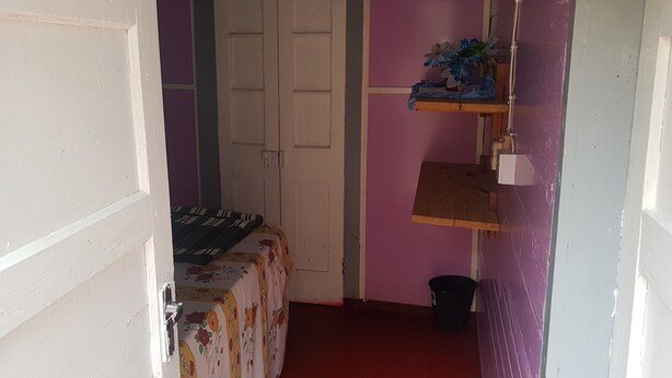 A narrow hallway with a bed, a door, and a small shelf with a bucket. - Jay's Guest House (Penlyne Castle)