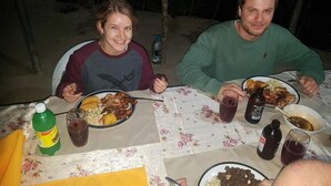 Dos personas cenando en una mesa al aire libre, con platos de comida, bebidas y una botella de refresco.