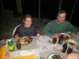 Two people dining at an outdoor table with plates of food, drinks, and a bottle of soda.