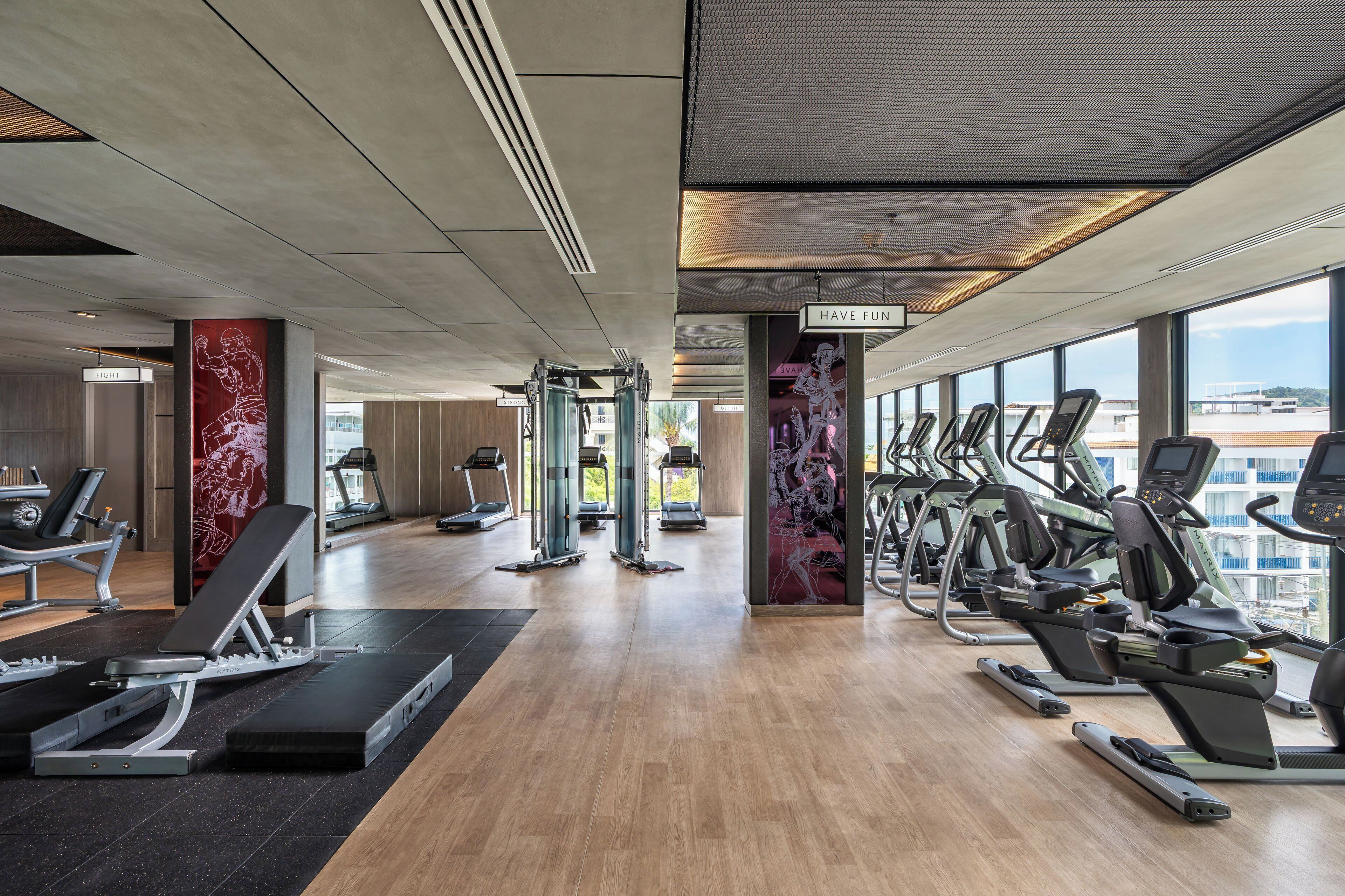 A modern gym with various exercise equipment, including treadmills and weight machines, and large windows offering views of the city.