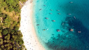 Vista aérea de una playa con aguas turquesas cristalinas, numerosos barcos y una orilla arenosa bordeada de palmeras.