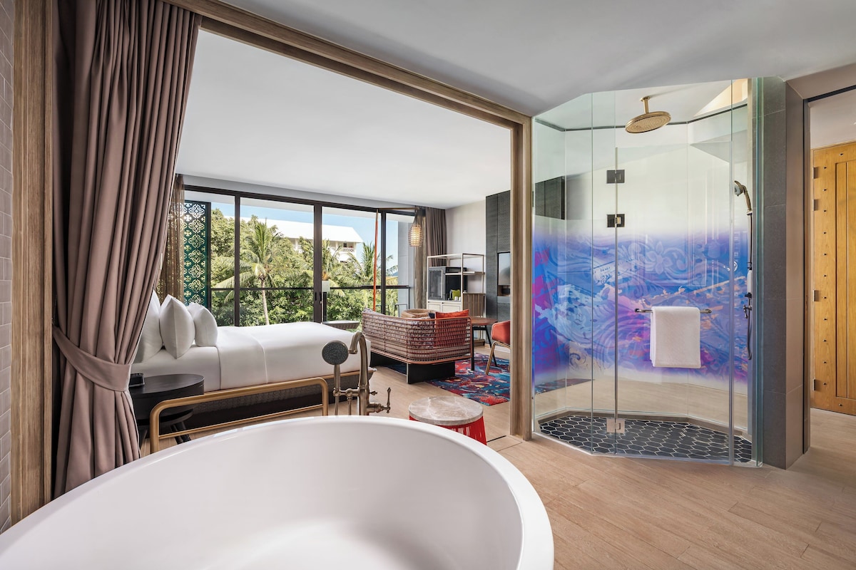 Habitación de hotel moderna con una bañera grande, ducha con cerramiento de vidrio, sofá y vistas al exterior.