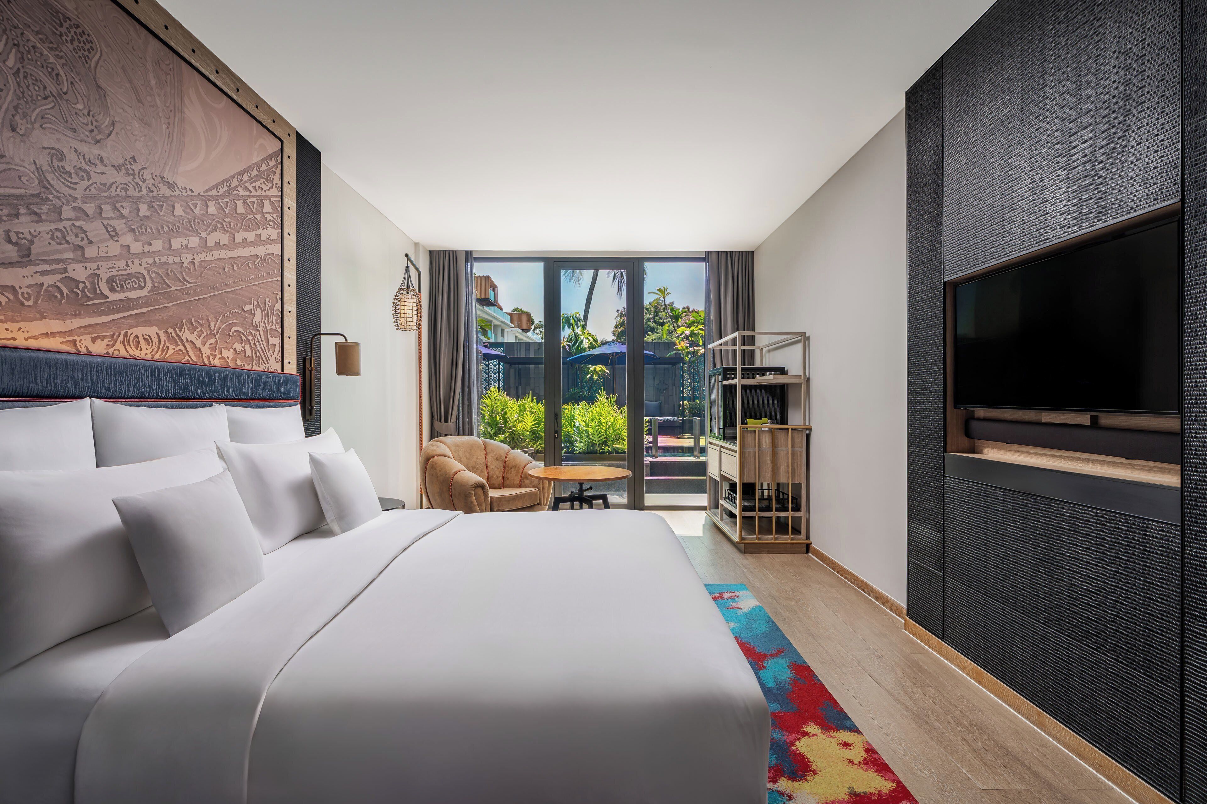 A modern hotel room with a large bed, a TV, a seating area with a chair and table, and a balcony with a view of greenery.