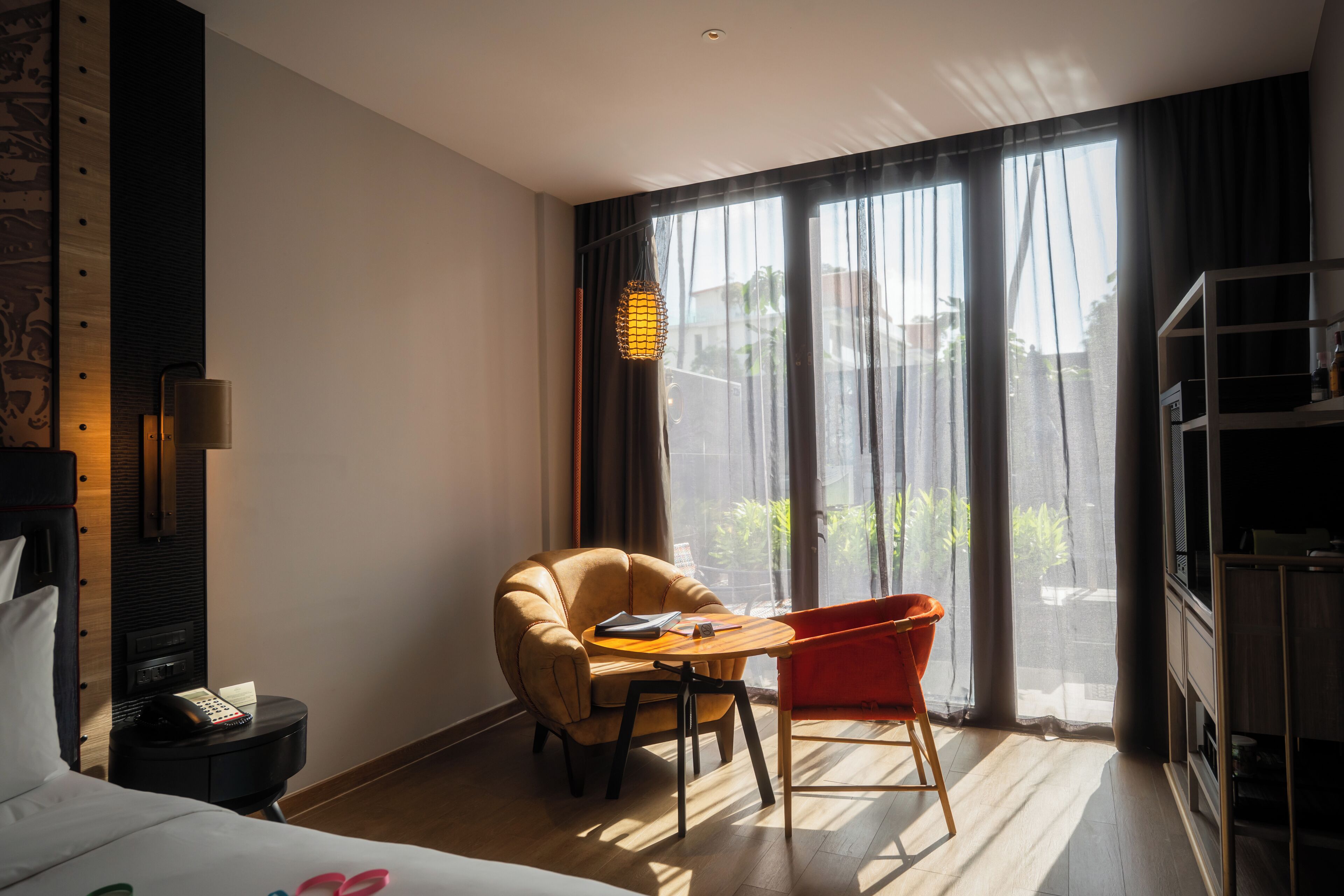A hotel room with a bed, a desk with a chair, a TV stand, and a large window with curtains.