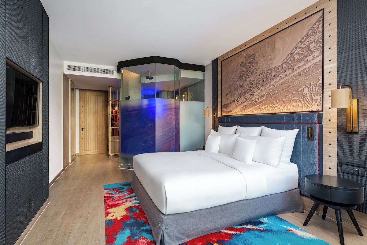 Una habitación de hotel moderna con una cama grande, una alfombra colorida, una obra de arte en la pared y una mesita de noche pequeña.
