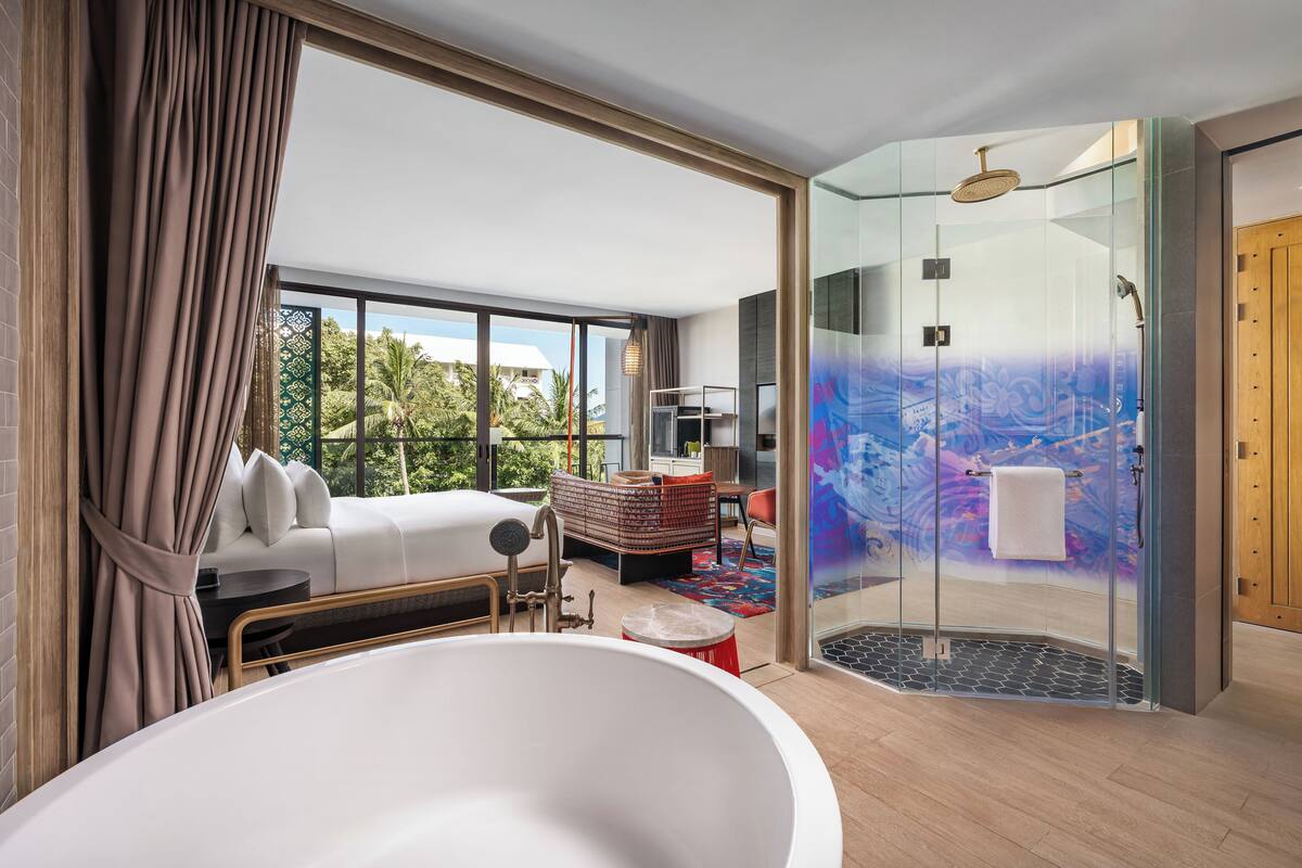 A modern hotel room with a large window, a glass-enclosed shower, a white bathtub, and a cozy seating area with a patterned sofa and a small table.