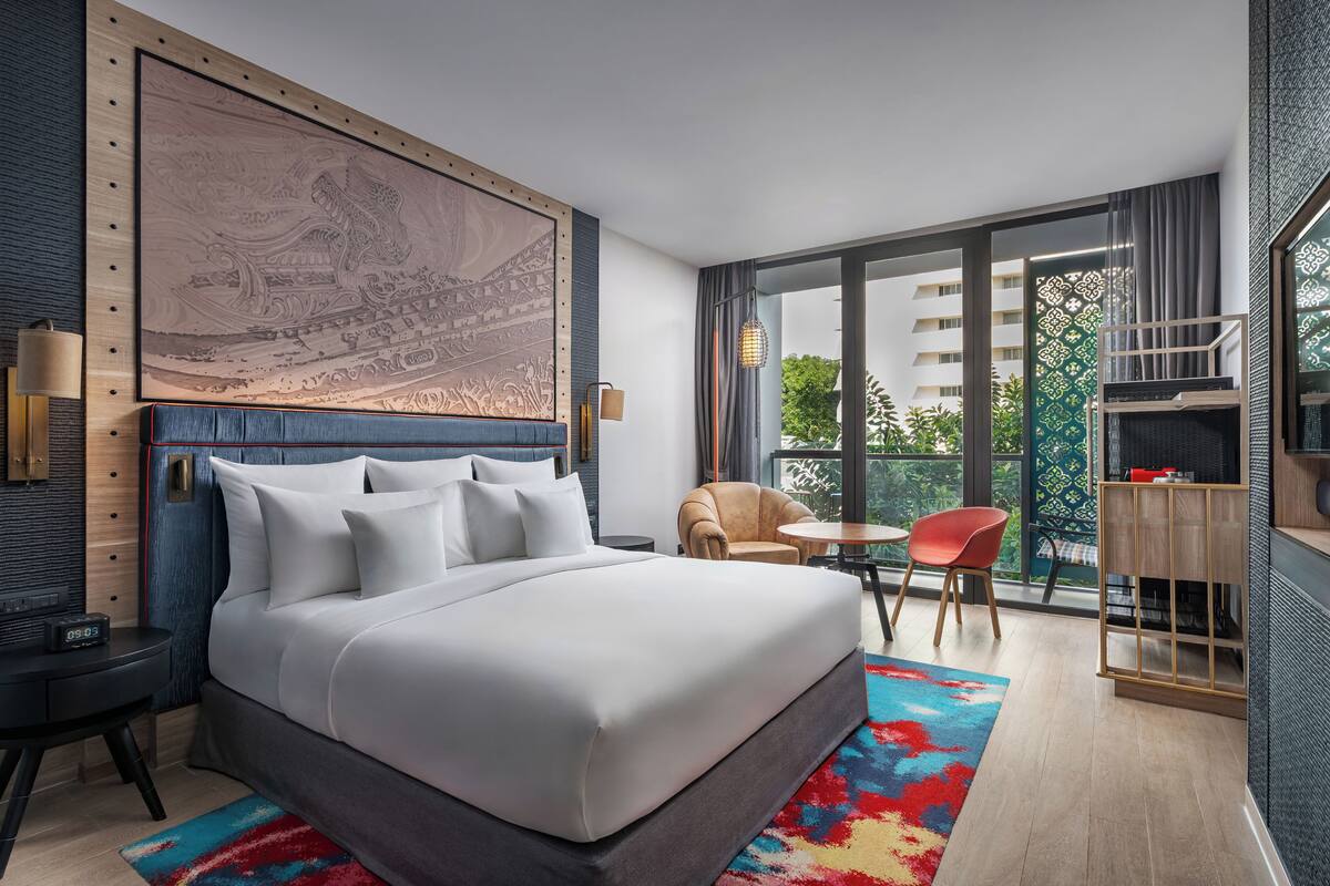 A modern hotel room with a large bed, a desk, a chair, and a balcony with a view of the city.