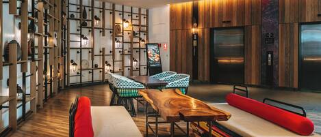 Un moderno vestíbulo de hotel con piso de madera, un área de estar central con una mesa y sillas de madera, y una gran pantalla de televisión montada en la pared.