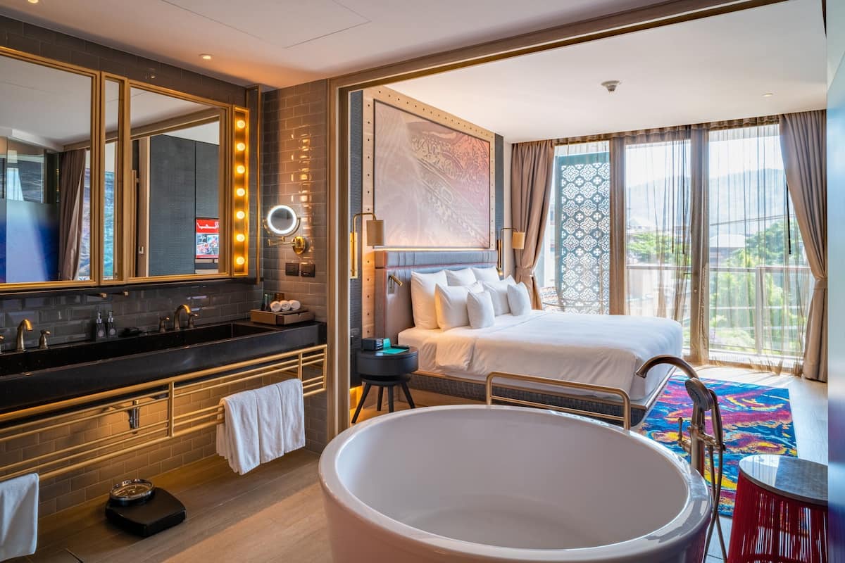 A modern hotel room with a large mirror, a bathtub, a double bed, and a balcony with a view.