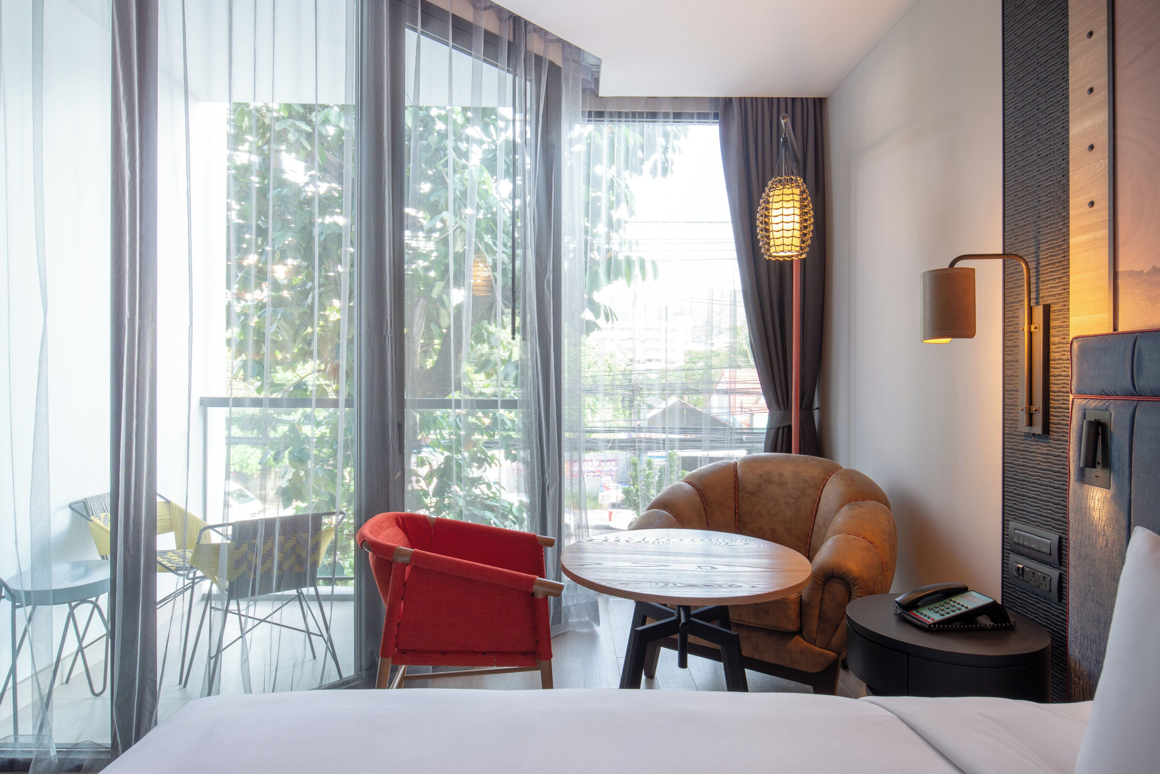 Une chambre d’hôtel avec un lit, un fauteuil rouge, une petite table ronde, un téléphone et une vue sur la verdure par la fenêtre.