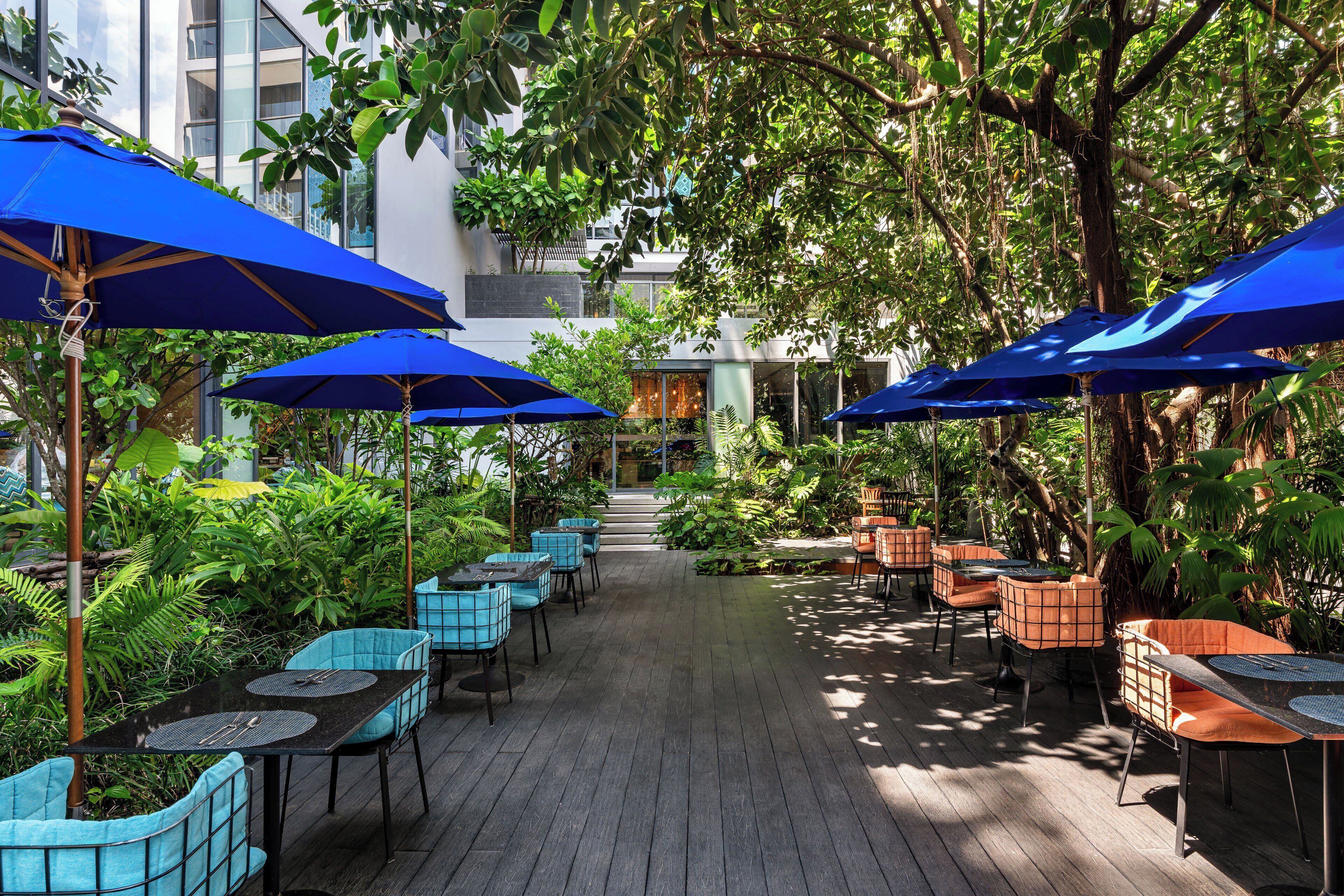 Un patio al aire libre con mesas y sillas, sombrillas azules y abundante vegetación.