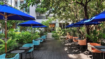 An outdoor patio with tables and chairs, blue umbrellas, and lush greenery.