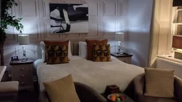 A hotel room with a large bed, two armchairs, a small table with fruit, a nightstand, a lamp, and a painting on the wall.