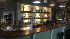 Zona de bar con estantes llenos de botellas de licores, mostrador con caja registradora y lámparas colgantes.