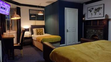 A compact hotel room with a bed, a desk, a chair, a small table, a lamp, a television, a door, and a fireplace.