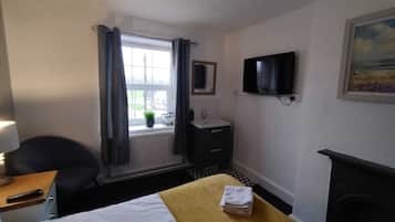 A hotel room with a bed, a desk with a lamp, a TV mounted on the wall, a small sink, and a window with curtains.