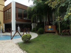 A modern building with a pool, a sign indicating 'Condomínio Enseada Praia do Forte', and lush greenery. - Apartamento Enseada Praia do Forte (Mata de São João)